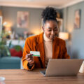 Woman shopping online on her laptop while holding a credit card at her desk.